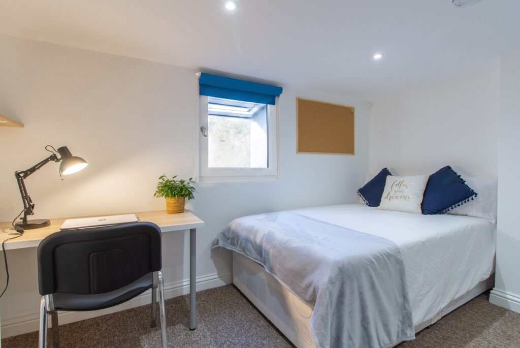 Bedroom 6 at 24 Granville Street, Cheltenham with a double bed, desk workspace, lamp, and skylight window with blue blind.